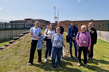 A group of 6 volunteers standing together on a grassy mound with buildings behind